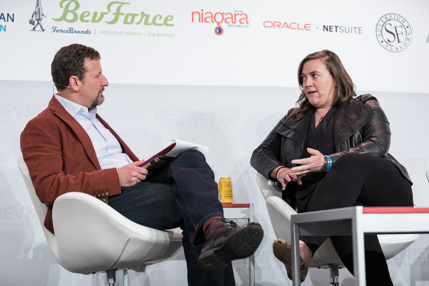 Interviewer in a rust jacket holding papers and a woman in a black leather jacket seated in white chairs, with a can on the table.