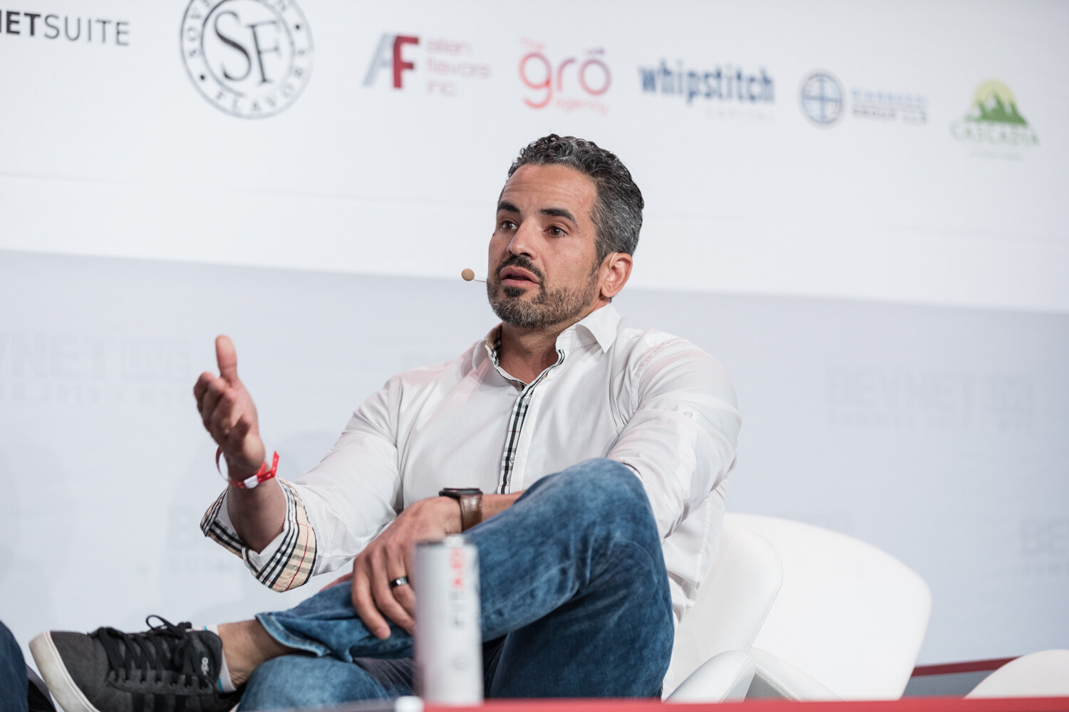 Seated man in a white shirt gesturing with an open hand, with a can on the table beside his knee.