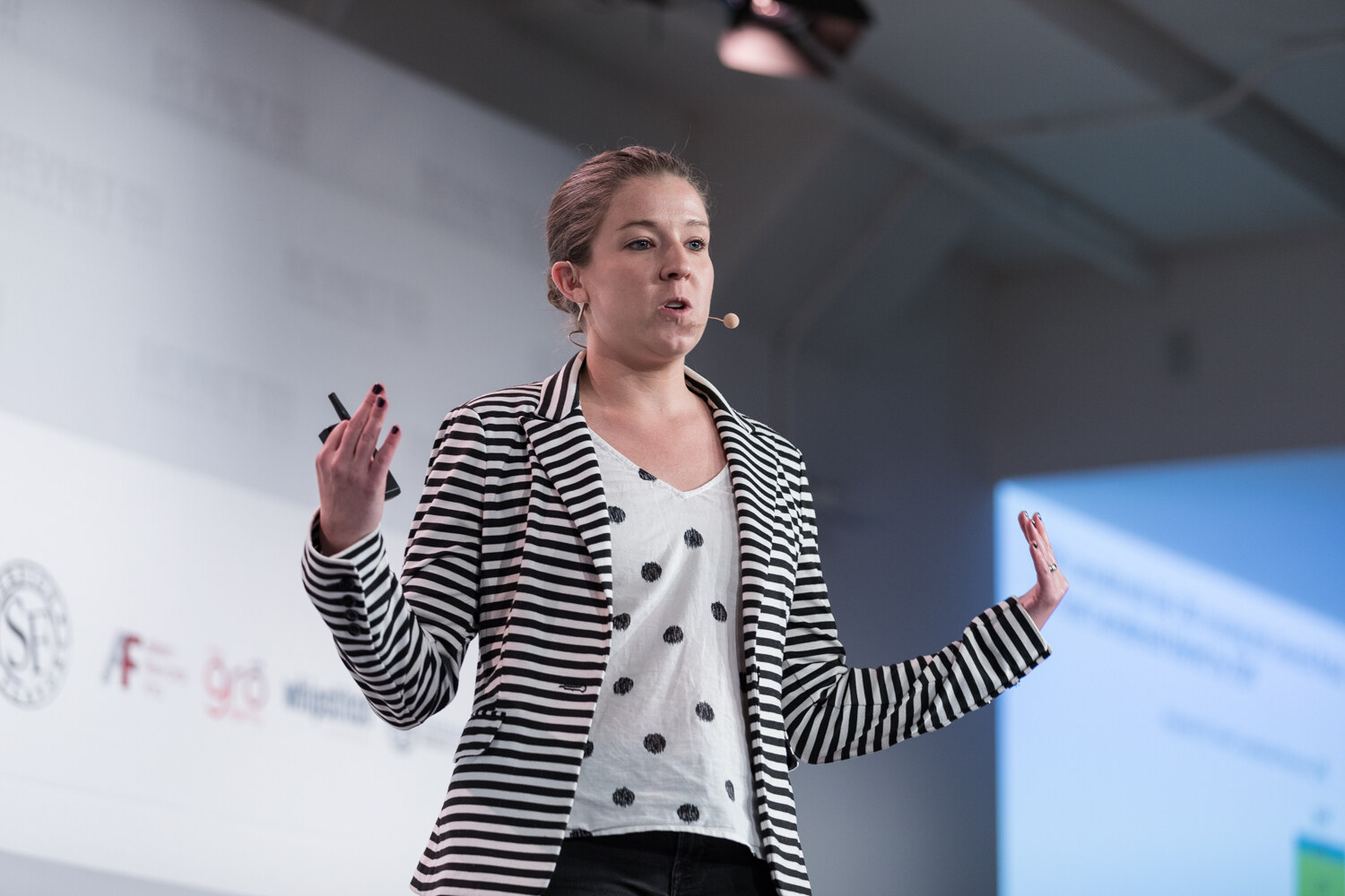 Woman in a striped blazer and polka-dot top with both hands raised in front of sponsor logos.