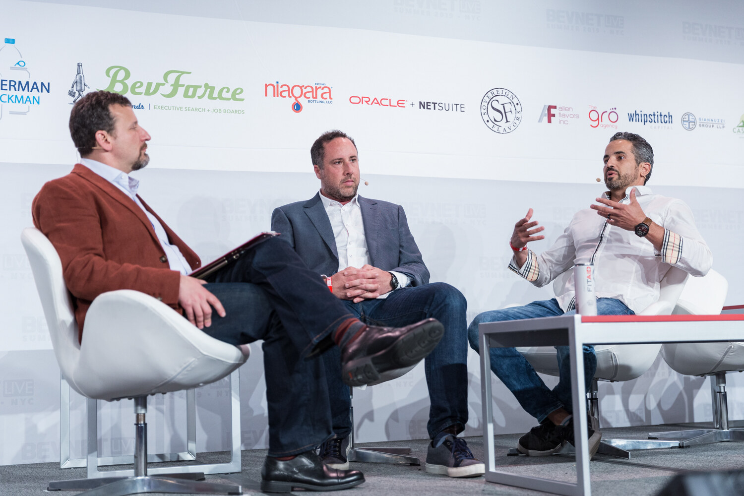 Interviewer in a rust jacket and two men seated in white chairs on the BevNET Live stage.