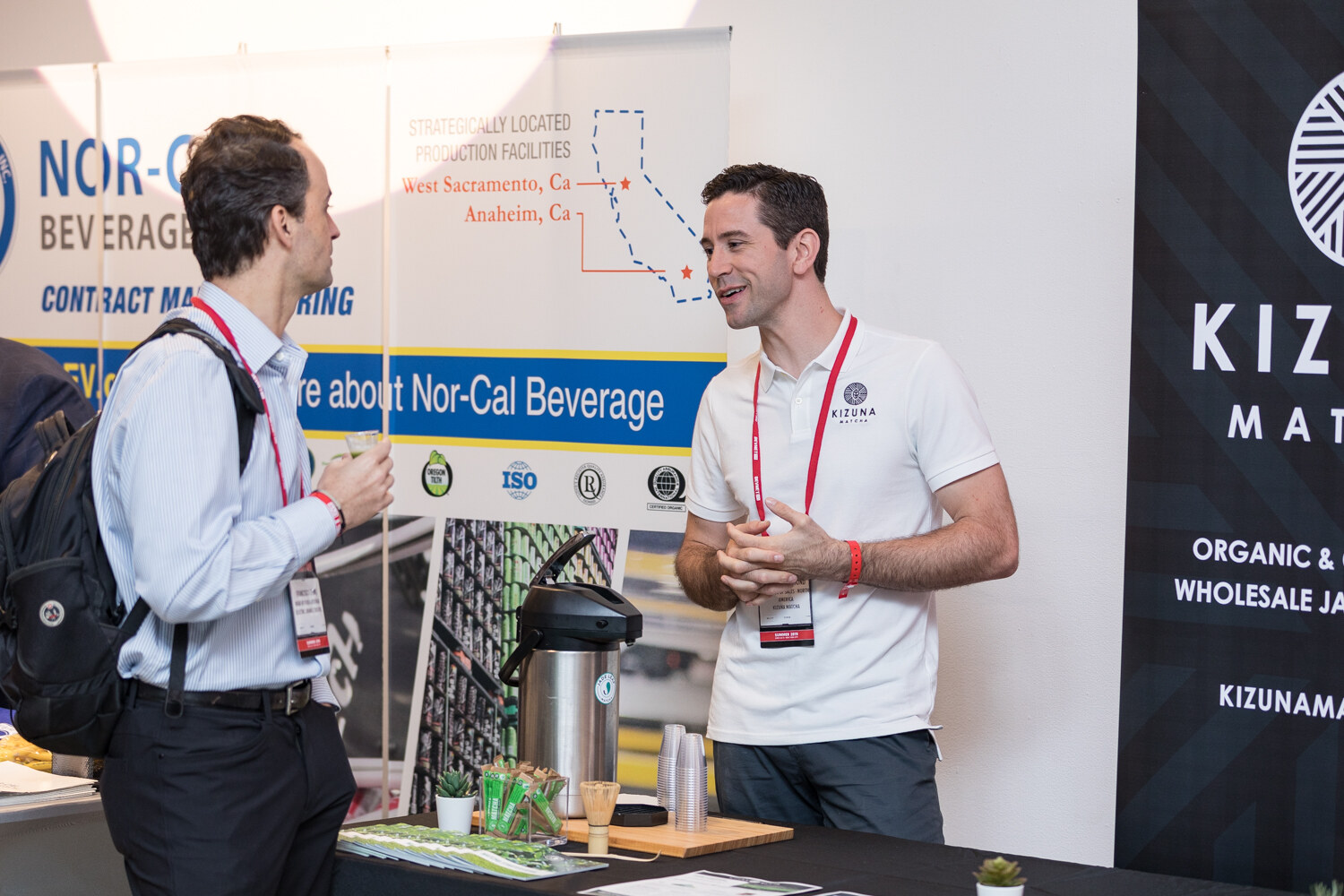 Man in a white polo talking with a visitor in front of a Nor-Cal Beverage Co. contract manufacturing banner and a Kizuna sign.