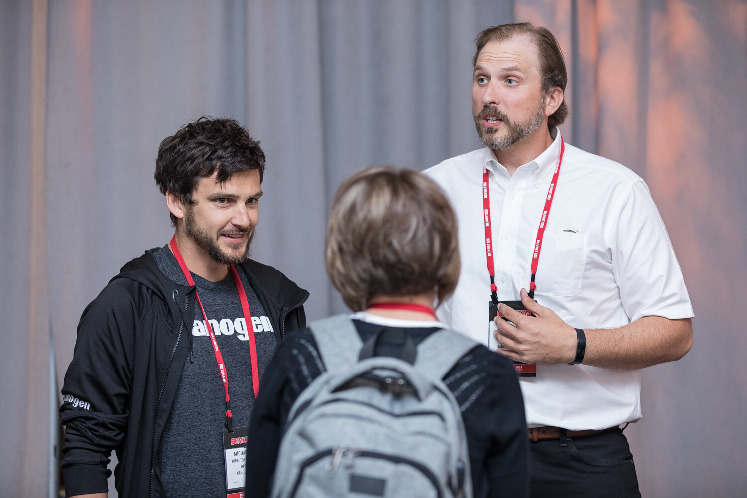 Man in a black Nanogen hoodie and a man in a white shirt talking with an attendee wearing a backpack.