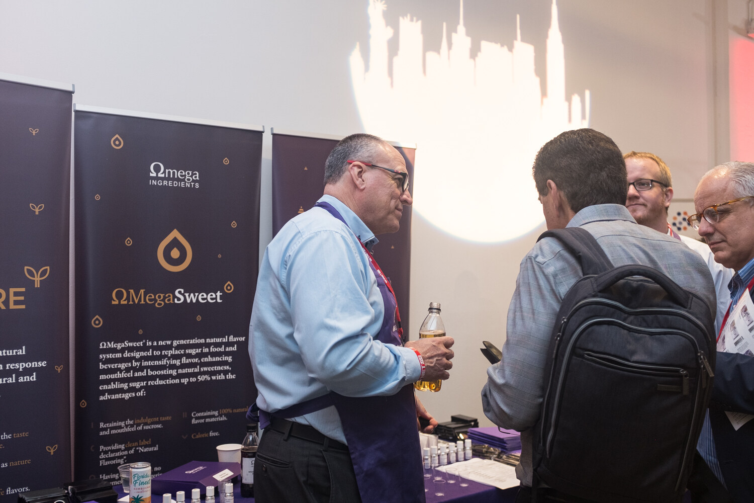 Exhibitor in a purple apron holding a sample bottle at the Omega Ingredients booth in front of an OmegaSweet banner.
