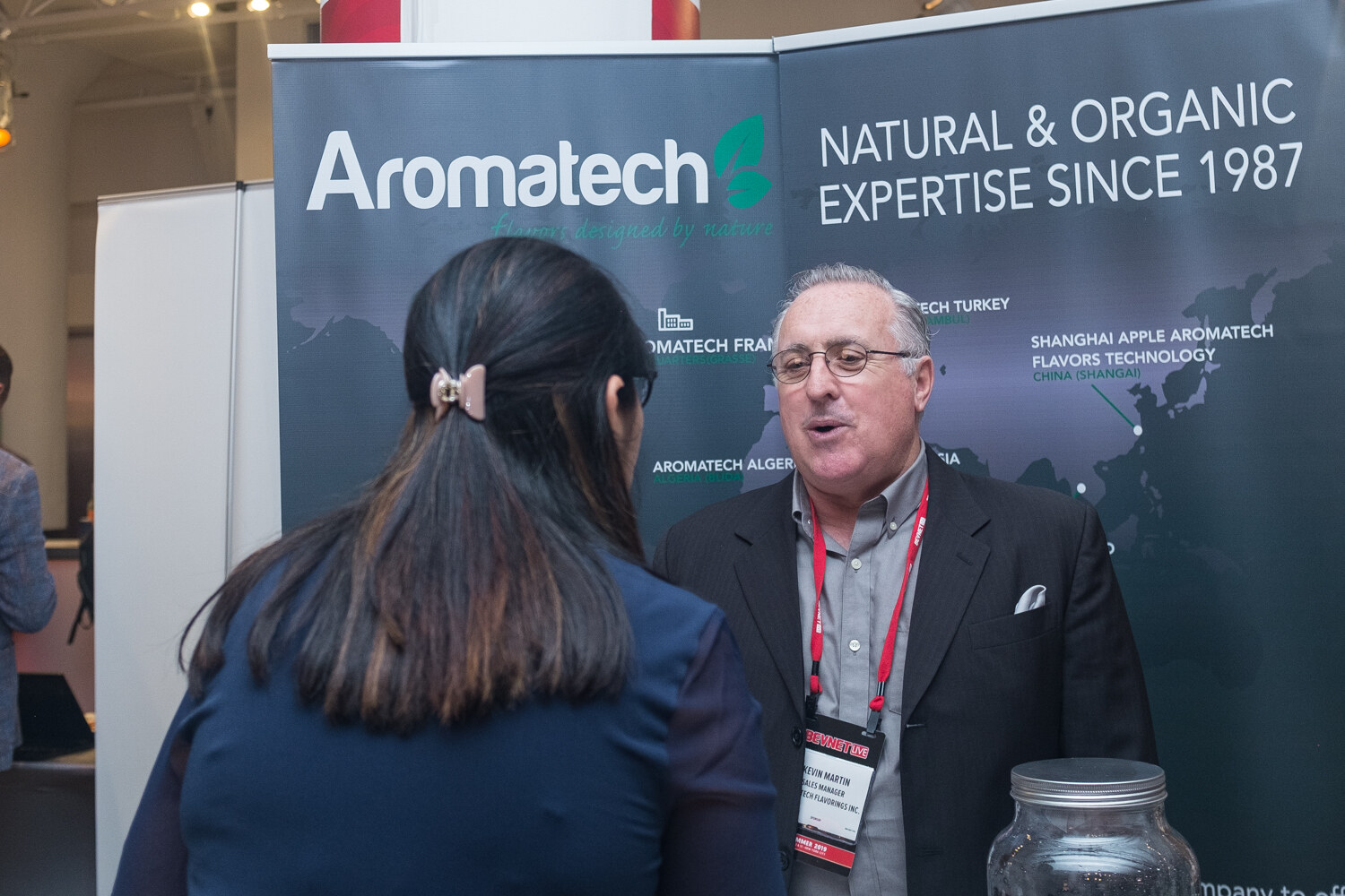 Man in a dark jacket talking with a woman in front of an Aromatech banner reading Natural and Organic Expertise Since 1987.
