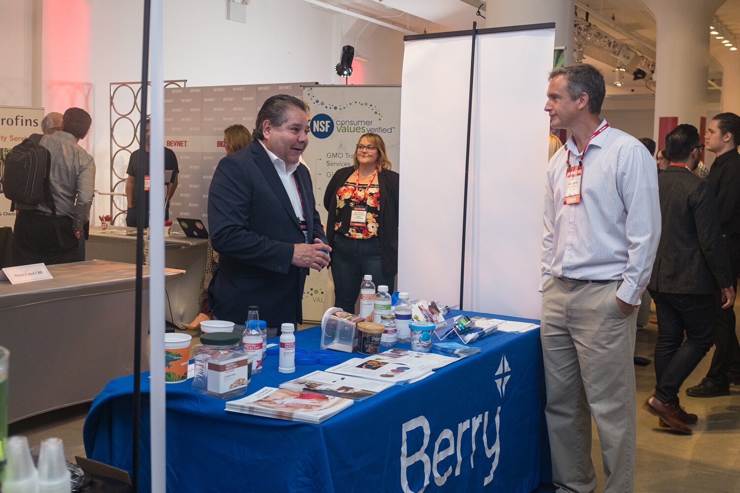 Two men and a woman talking at a booth with a blue Berry tablecloth, brochures and sample cups.