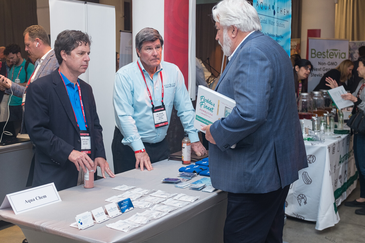 Three men talking at an Aqua Chem table covered with sample packets, with a Bestevia banner behind them.