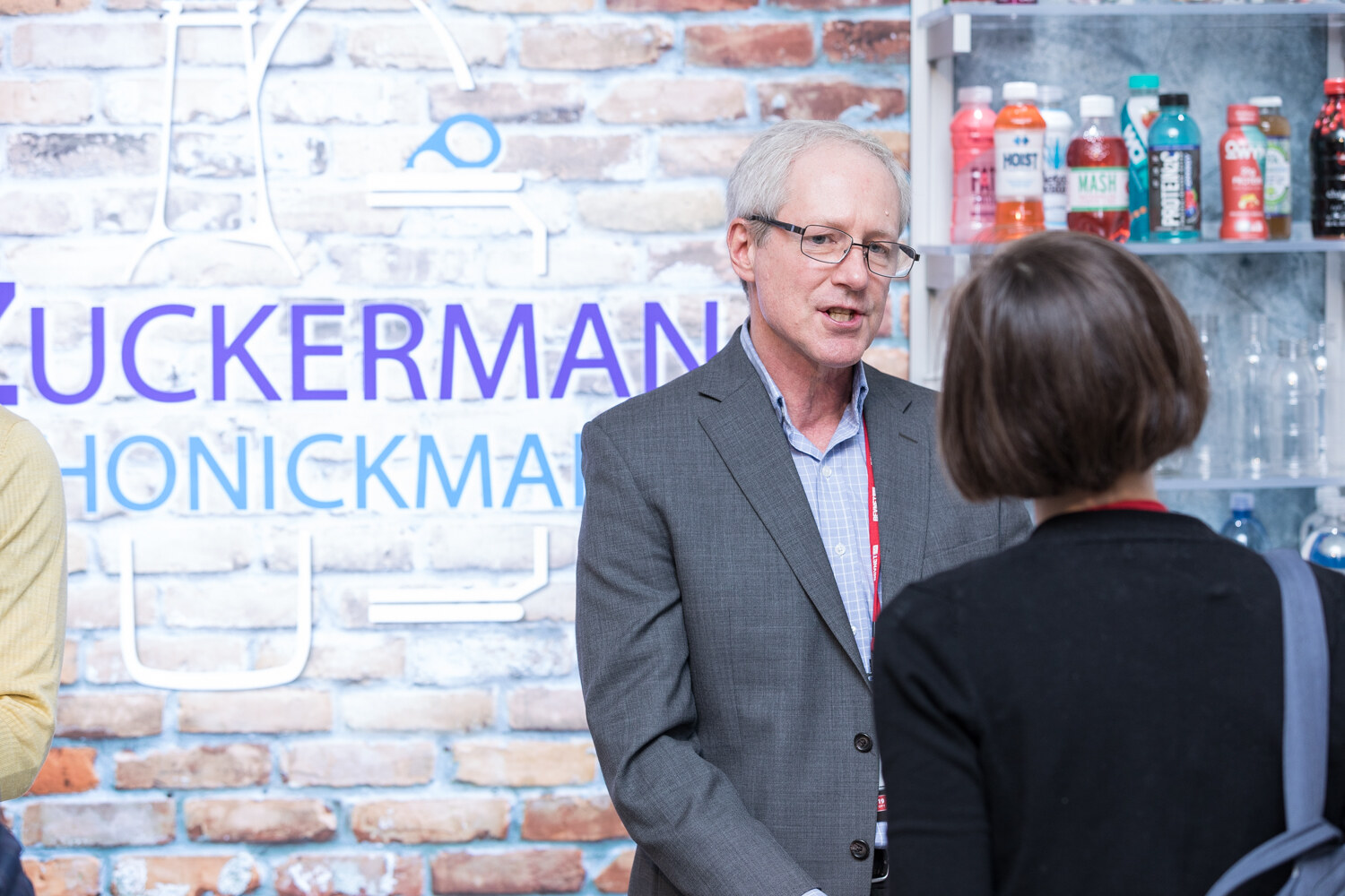 Man in a gray blazer talking with an attendee in front of a brick wall with a Zuckerman Honickman sign.