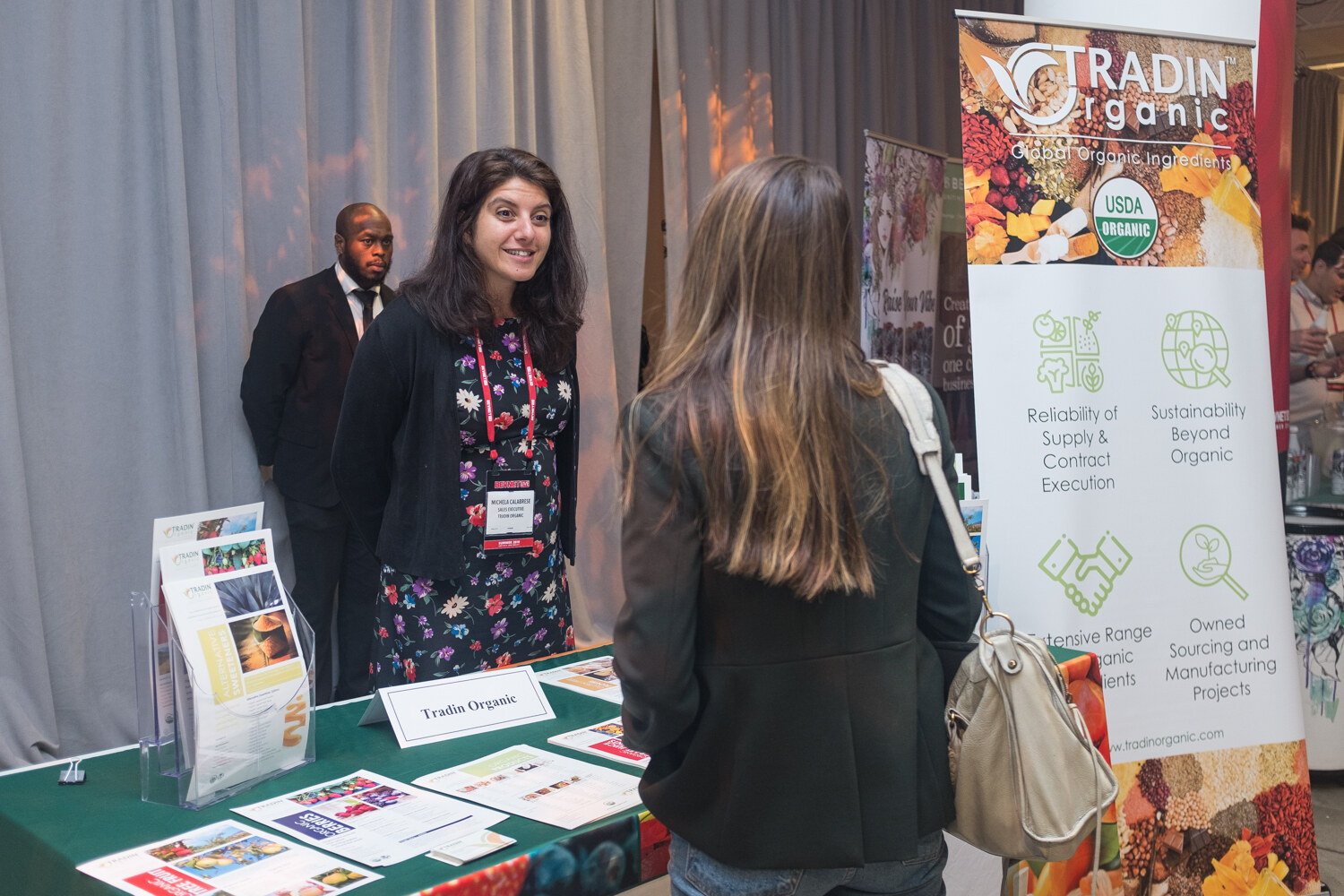 Woman exhibitor in a dark cardigan at the Tradin Organic booth beside a USDA Organic banner as a visitor stops to talk.