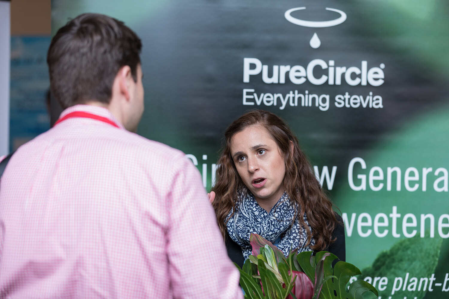 Woman in a blue scarf speaking with a man in a pink shirt in front of a PureCircle Everything stevia banner.