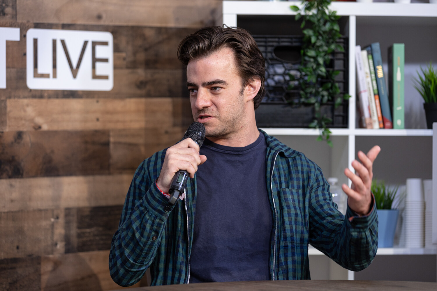 Man in a green plaid overshirt and navy t-shirt gesturing with an open hand while speaking into a microphone.