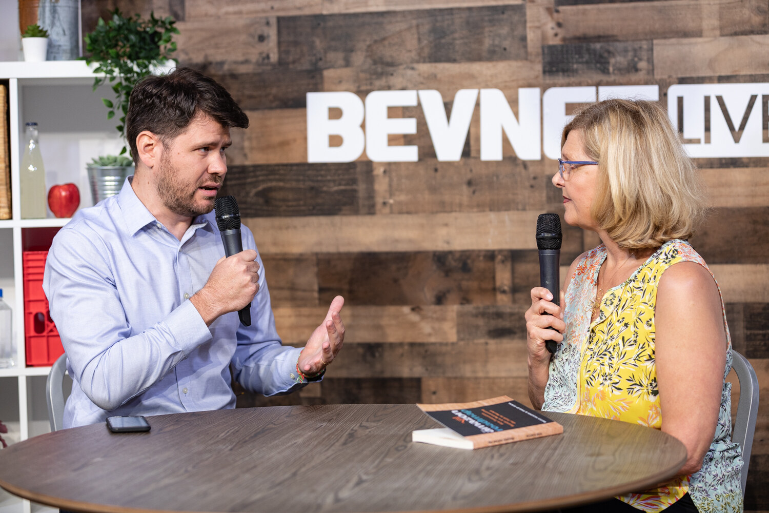 Man in a light blue shirt and a woman in a yellow floral top in conversation at the livestream table, with a book on it.