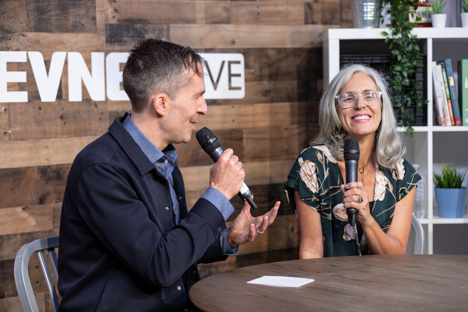 Man in a navy overshirt talking with a smiling woman with gray hair in a floral top at the livestream table.