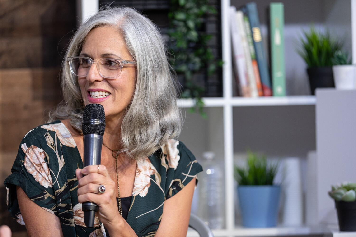 Smiling woman with gray hair and clear glasses speaking into a microphone.