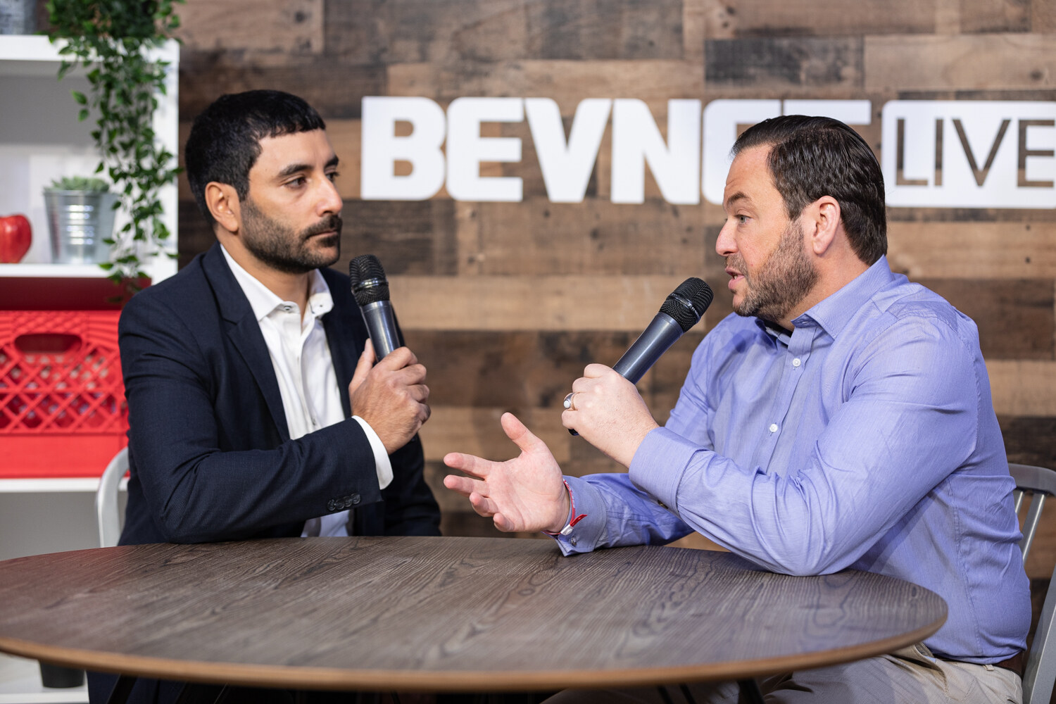 Man in a dark suit and a man in a lavender dress shirt gesturing with an open hand at the livestream table in front of the BevNET Live wall.