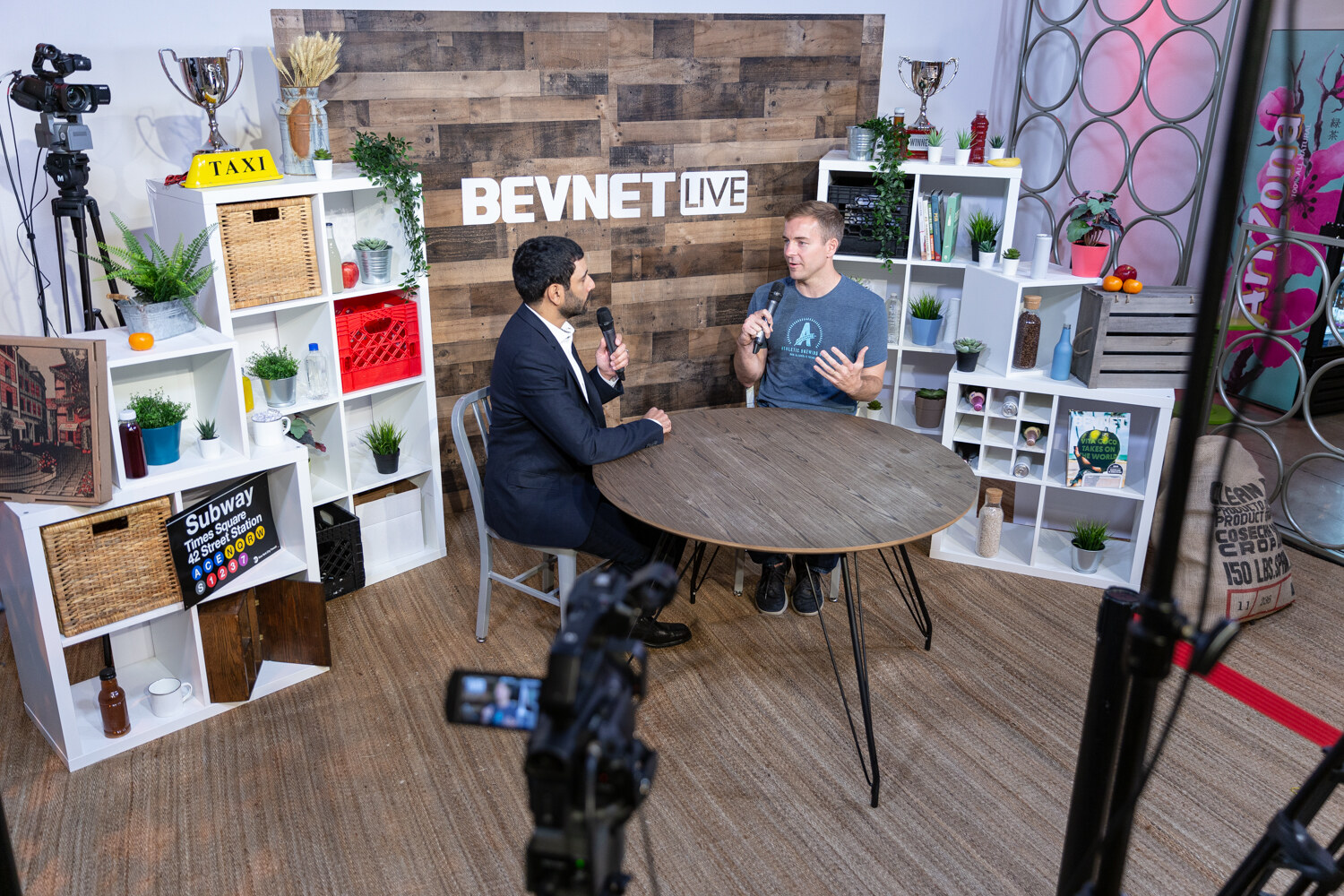 Wide view of the Taste Radio livestream set with two men talking at a round table in front of a wood-plank BevNET Live wall, with camera equipment in the foreground.
