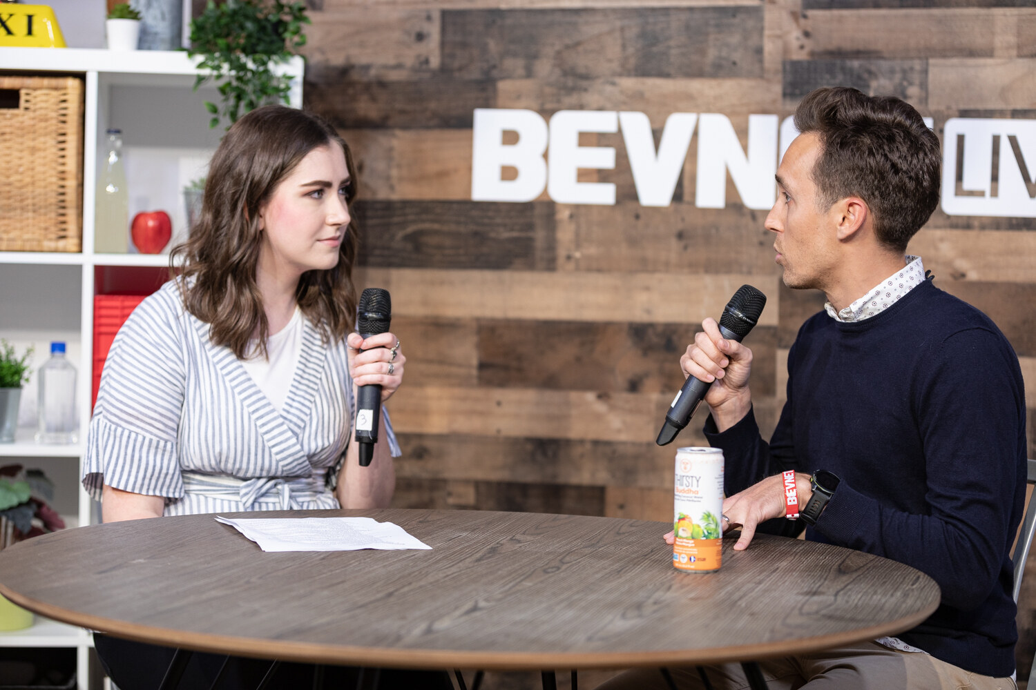Woman in a striped wrap dress interviewing a man in a navy sweater at the livestream table, with a Thirsty Buddha can between them.