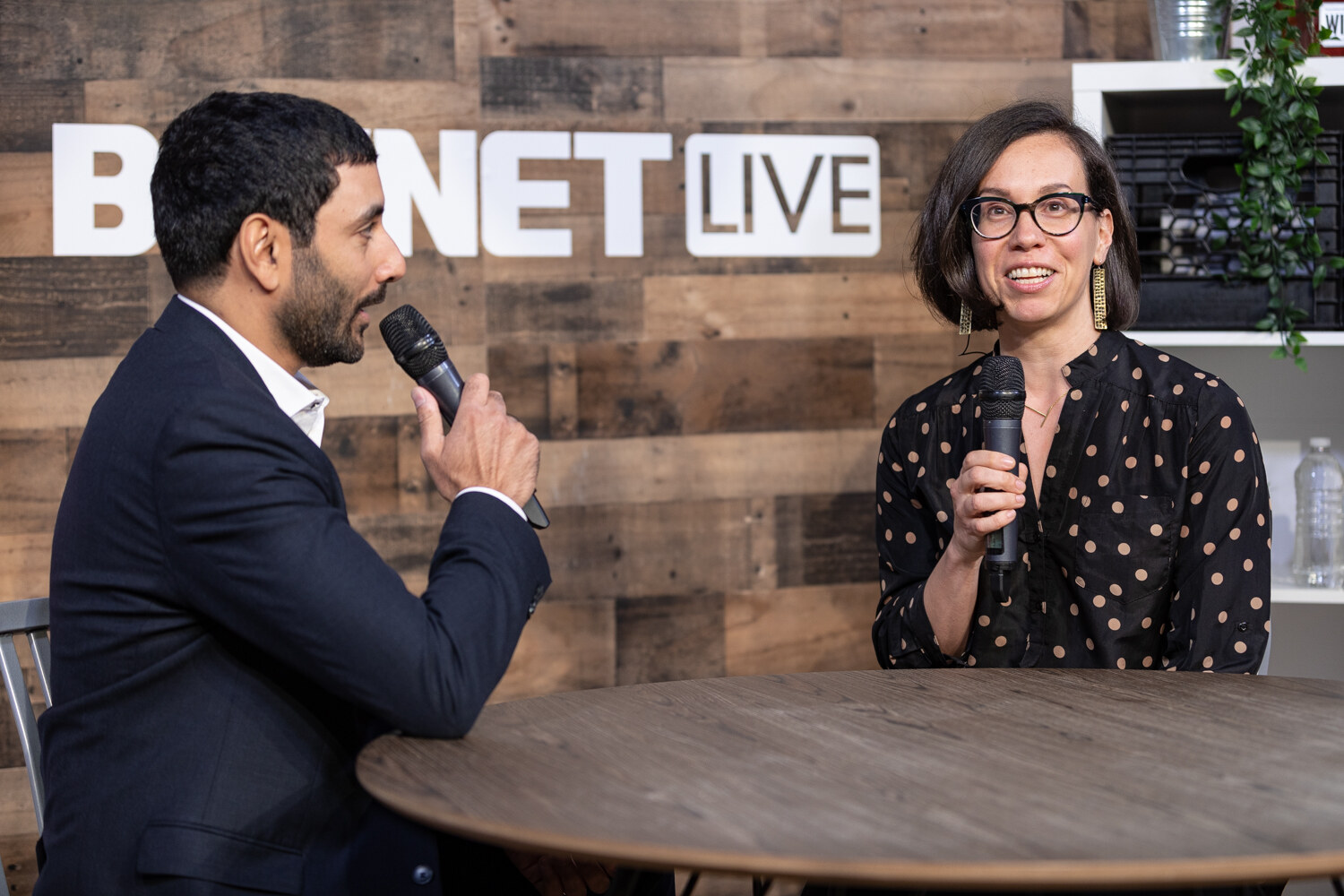 Man in a navy suit facing a woman in a black polka-dot blouse and round glasses at the livestream table.