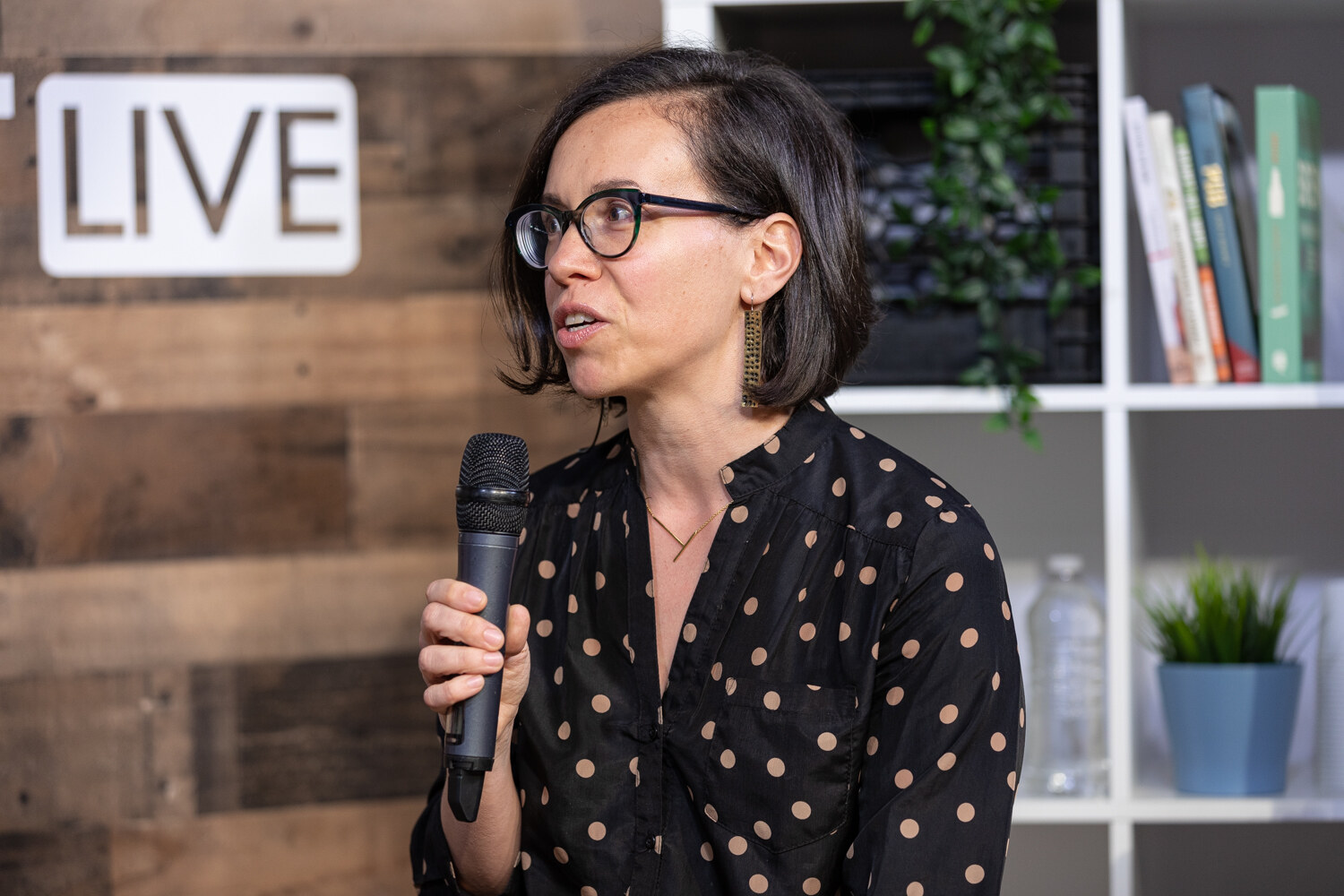 Woman in a black polka-dot blouse and round glasses speaking into a microphone at the livestream set.