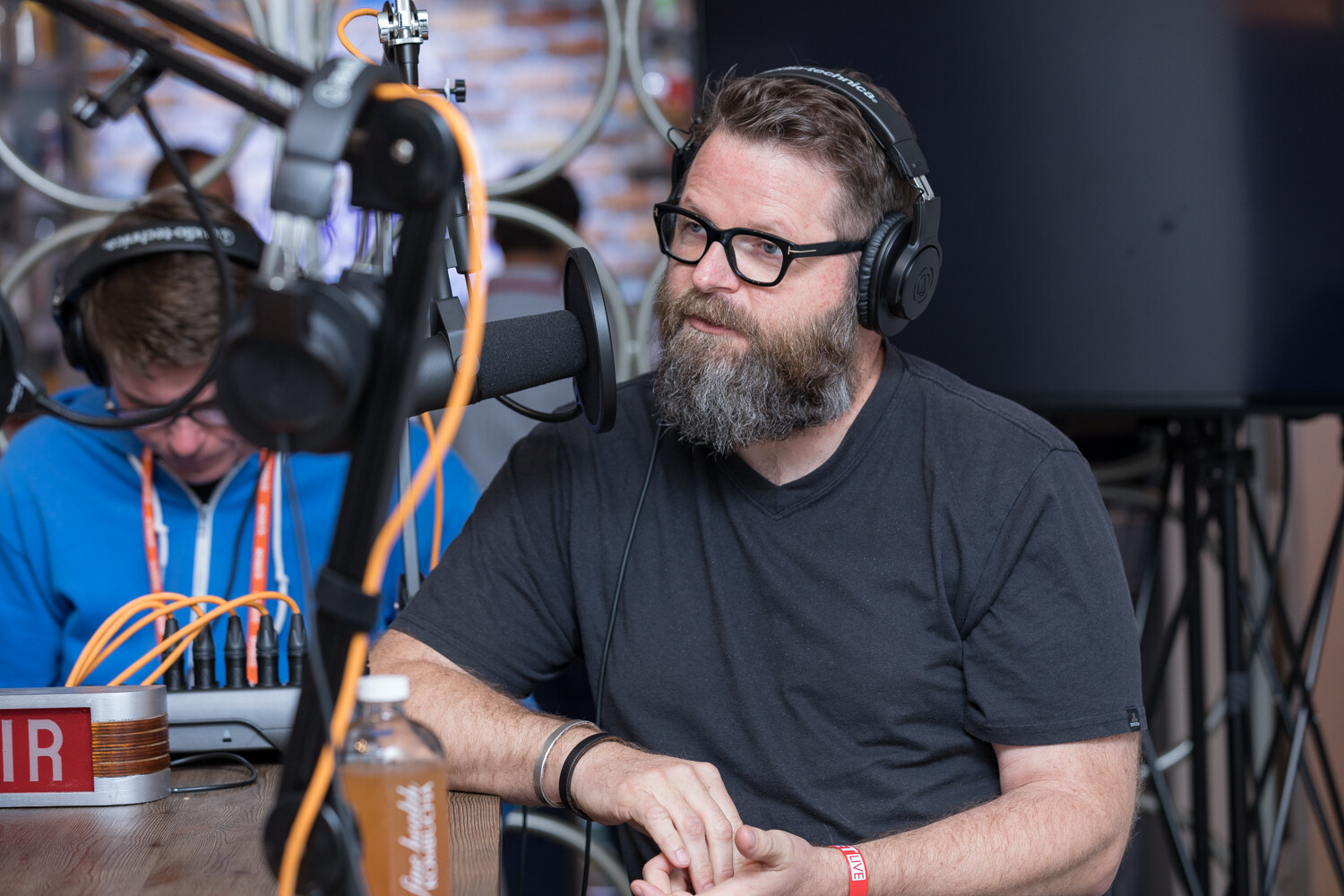 Bearded man with glasses and headphones seated at a microphone at the Taste Radio livestream table.