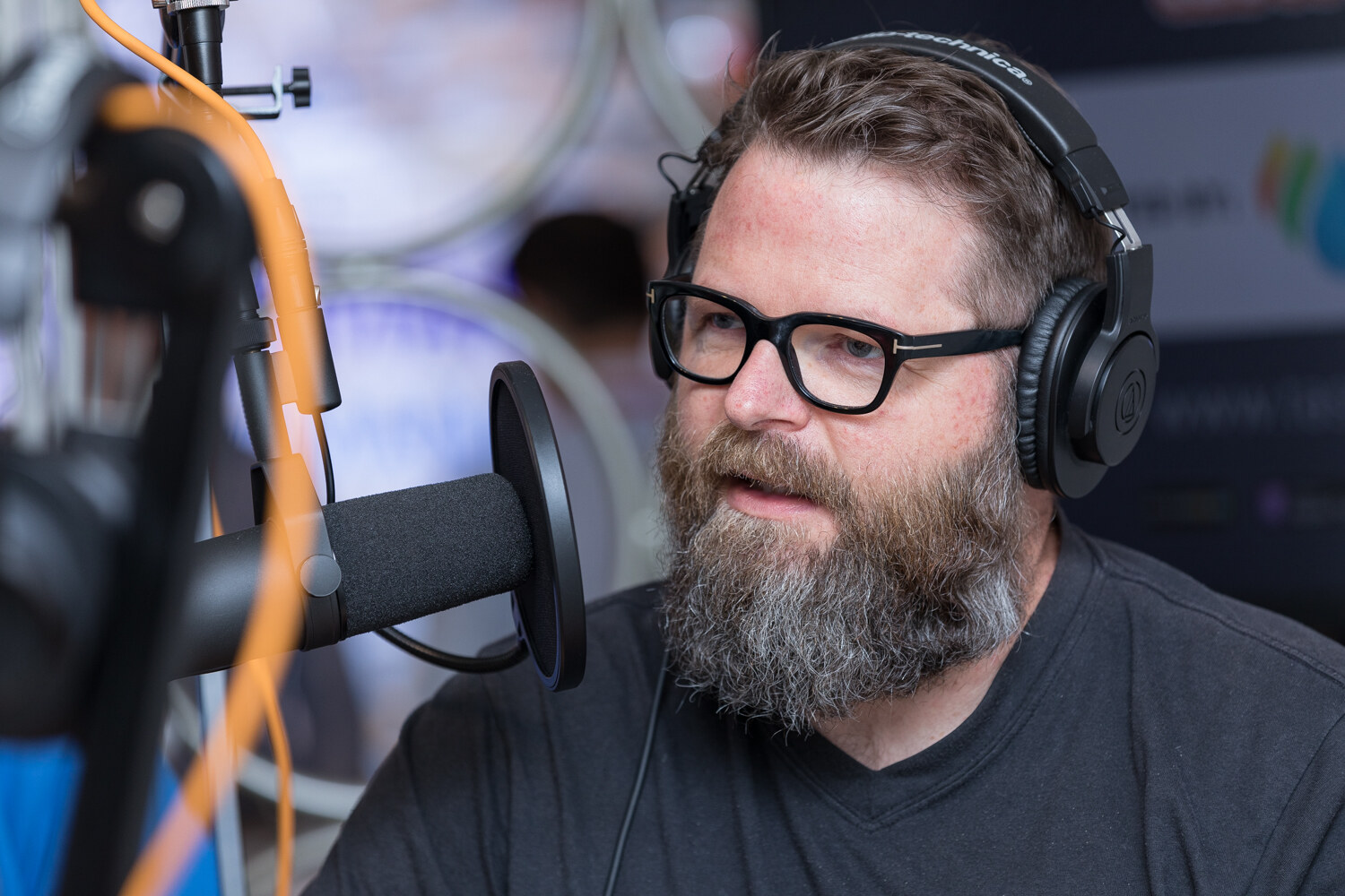 Close view of a bearded man in glasses and headphones speaking into a microphone at the Taste Radio livestream table.