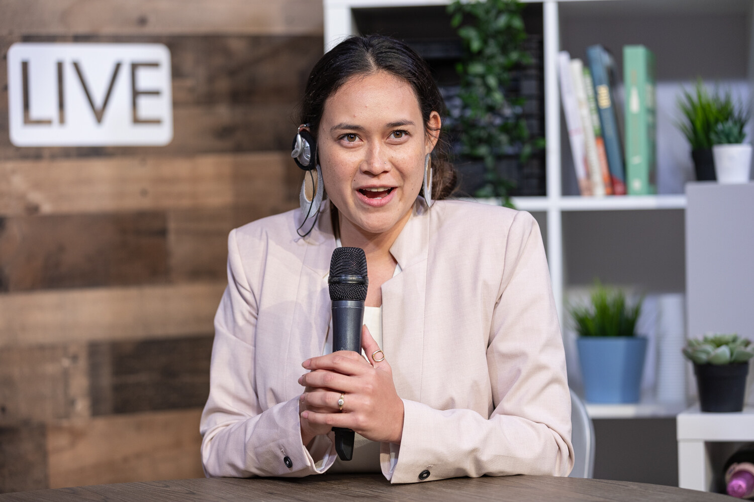 Woman in a cream blazer and statement earrings speaking into a microphone at the livestream set.