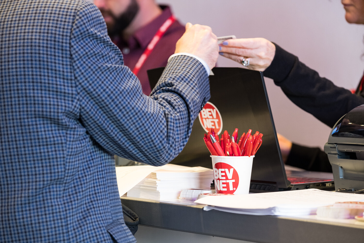 Close view of a red lanyard being handed across the registration counter beside a BevNET cup of red pens.
