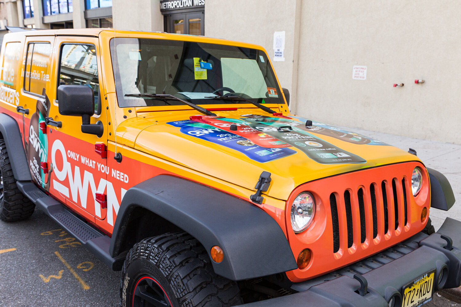 Front of a yellow and orange OWYN-wrapped Jeep parked on the street beside the Metropolitan West entrance.