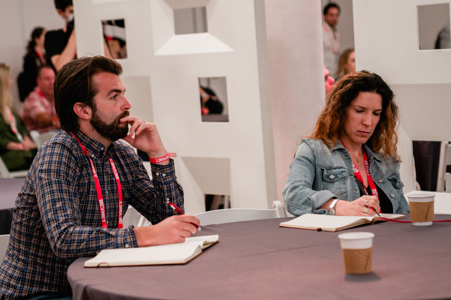 A man and a woman in lanyards sit at a round table with notebooks and coffee cups, writing.