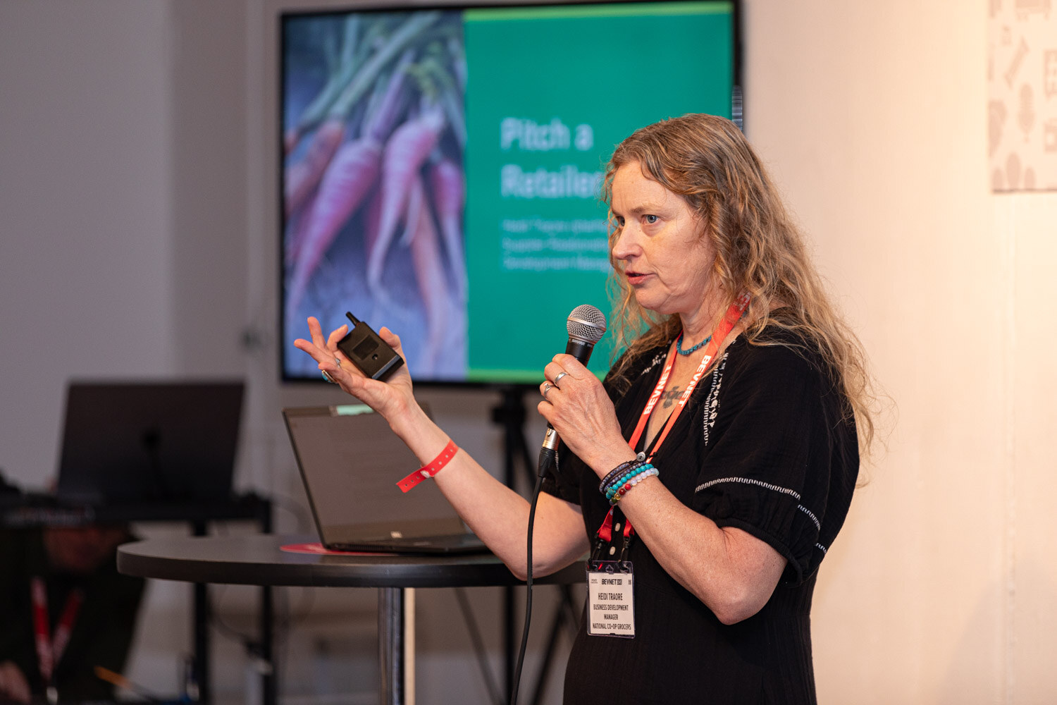 A woman with a microphone and a clicker speaks in front of a screen reading Pitch a Retailer.