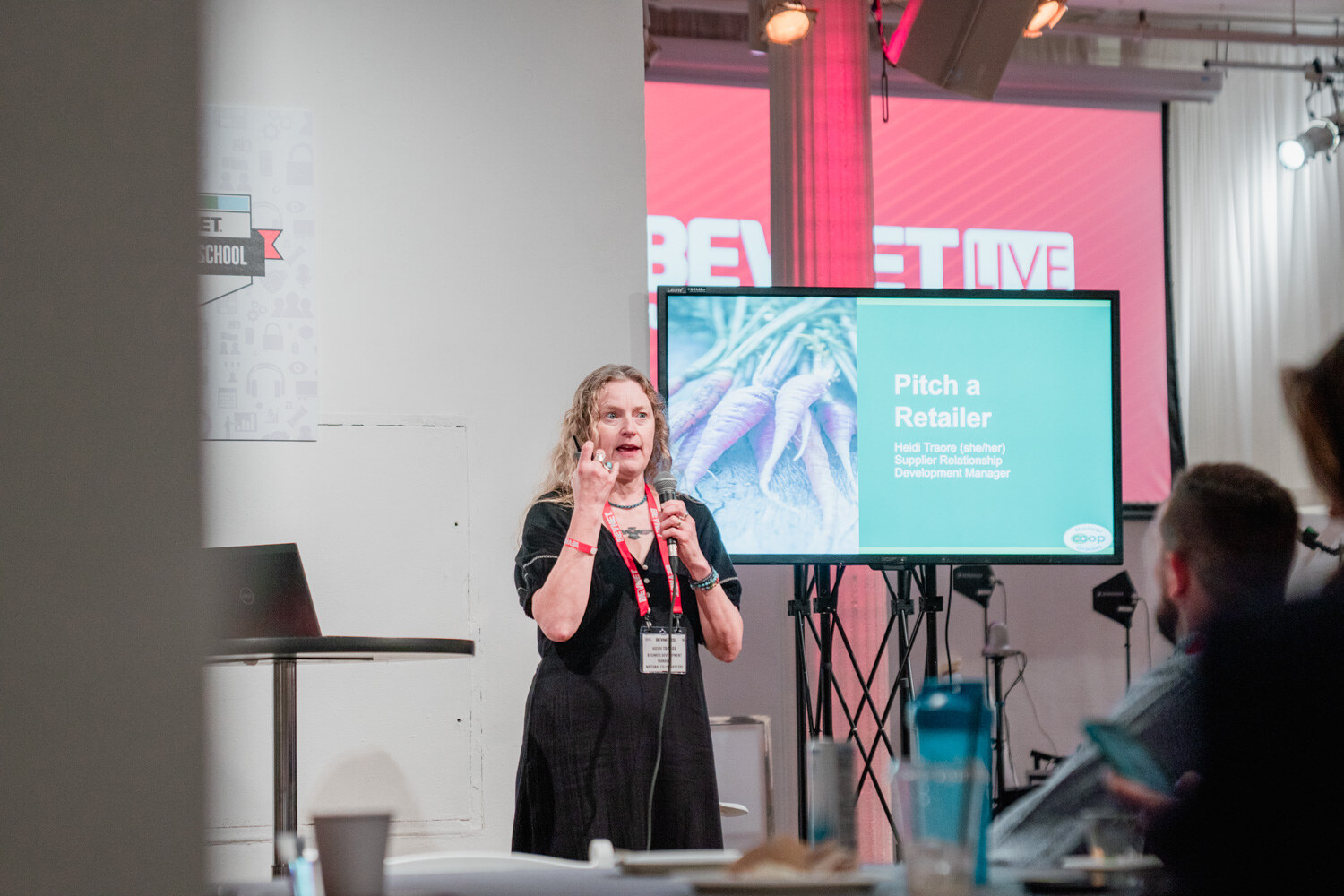 A woman presents next to a screen titled Pitch a Retailer, with attendees in the foreground and a BevNET Beverage School sign behind.