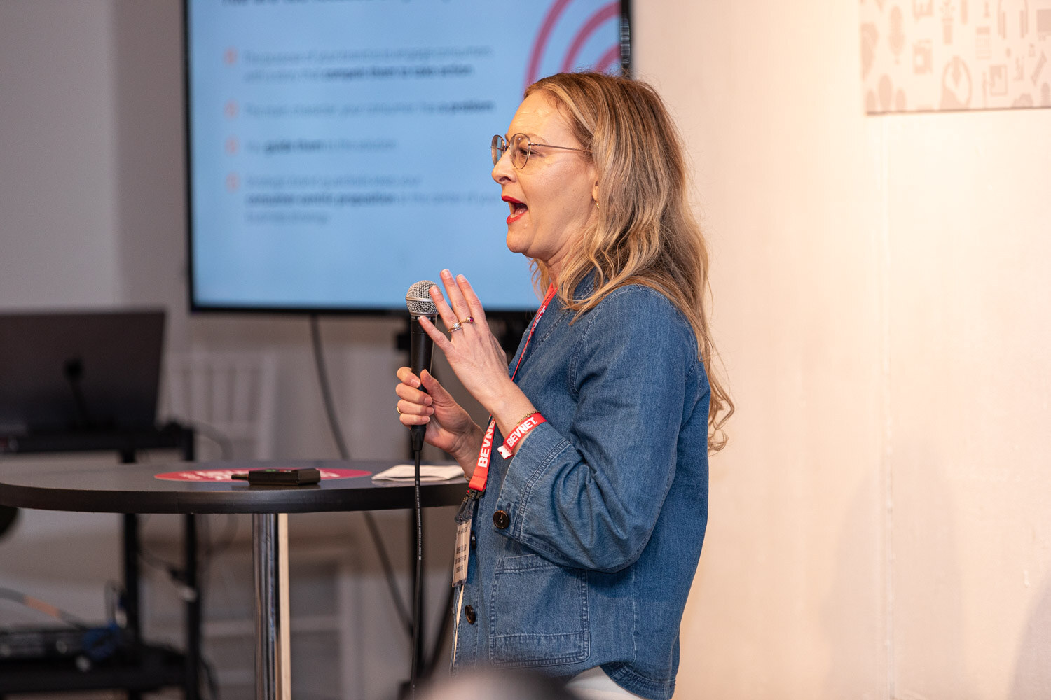 A woman holding a microphone gestures while speaking in front of a blurred presentation slide.