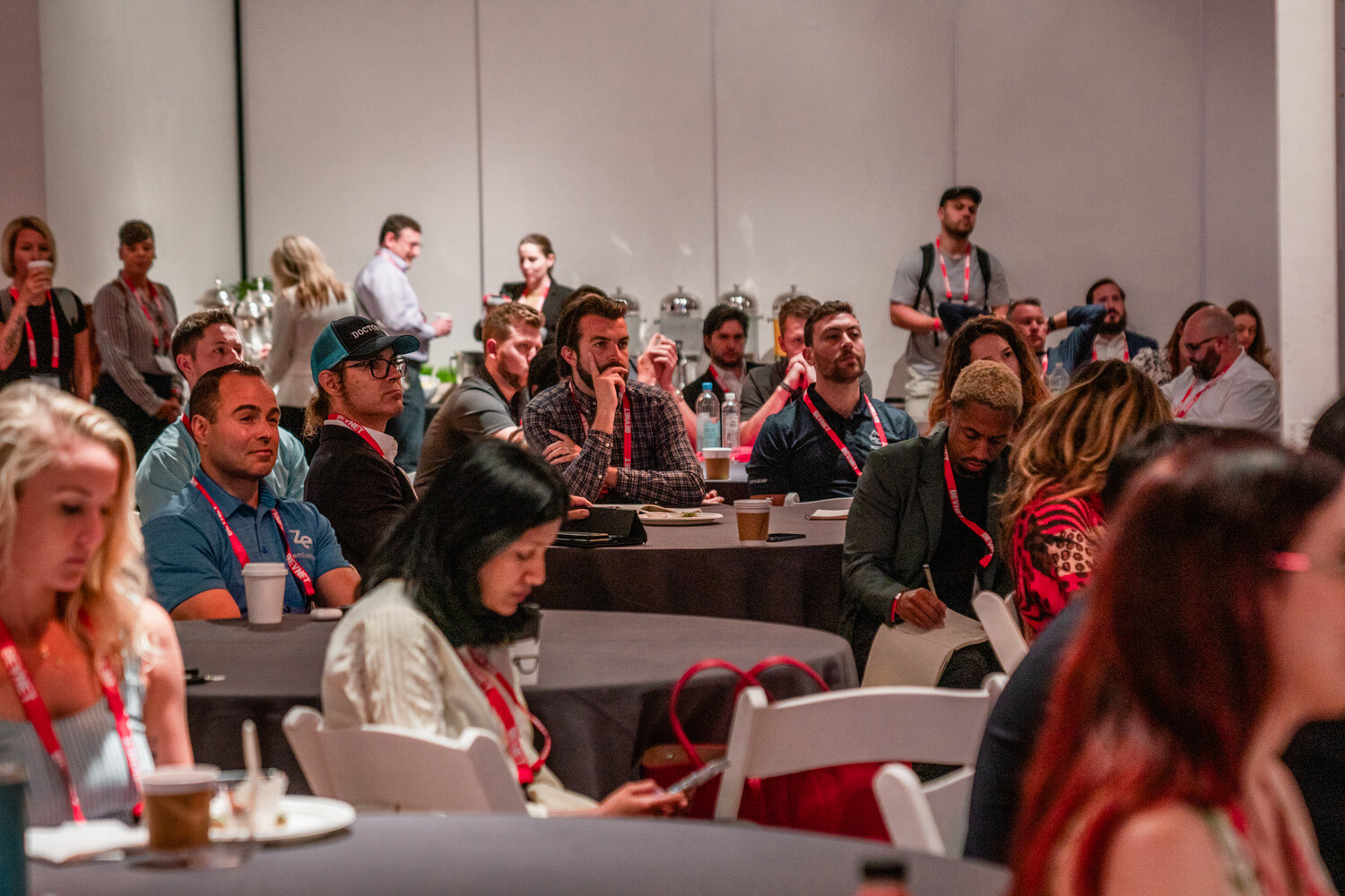 A crowded room of attendees in lanyards seated at round tables, some holding coffee and notes.