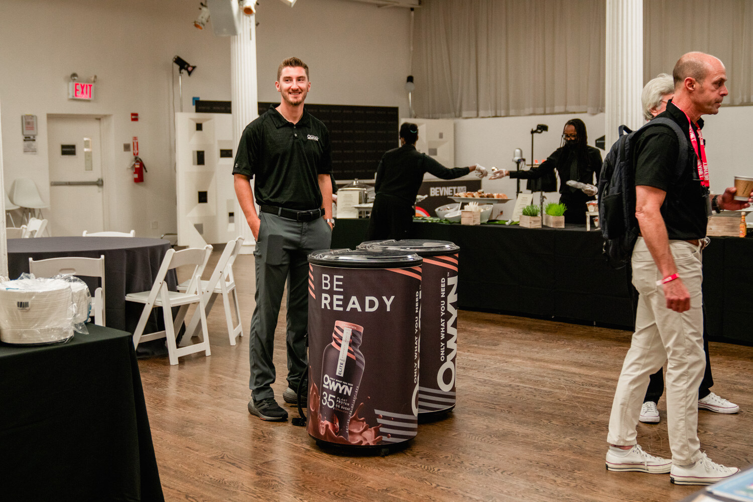 Two men stand near a black Owyn Be Ready cooler in a large room with a breakfast buffet in the background.