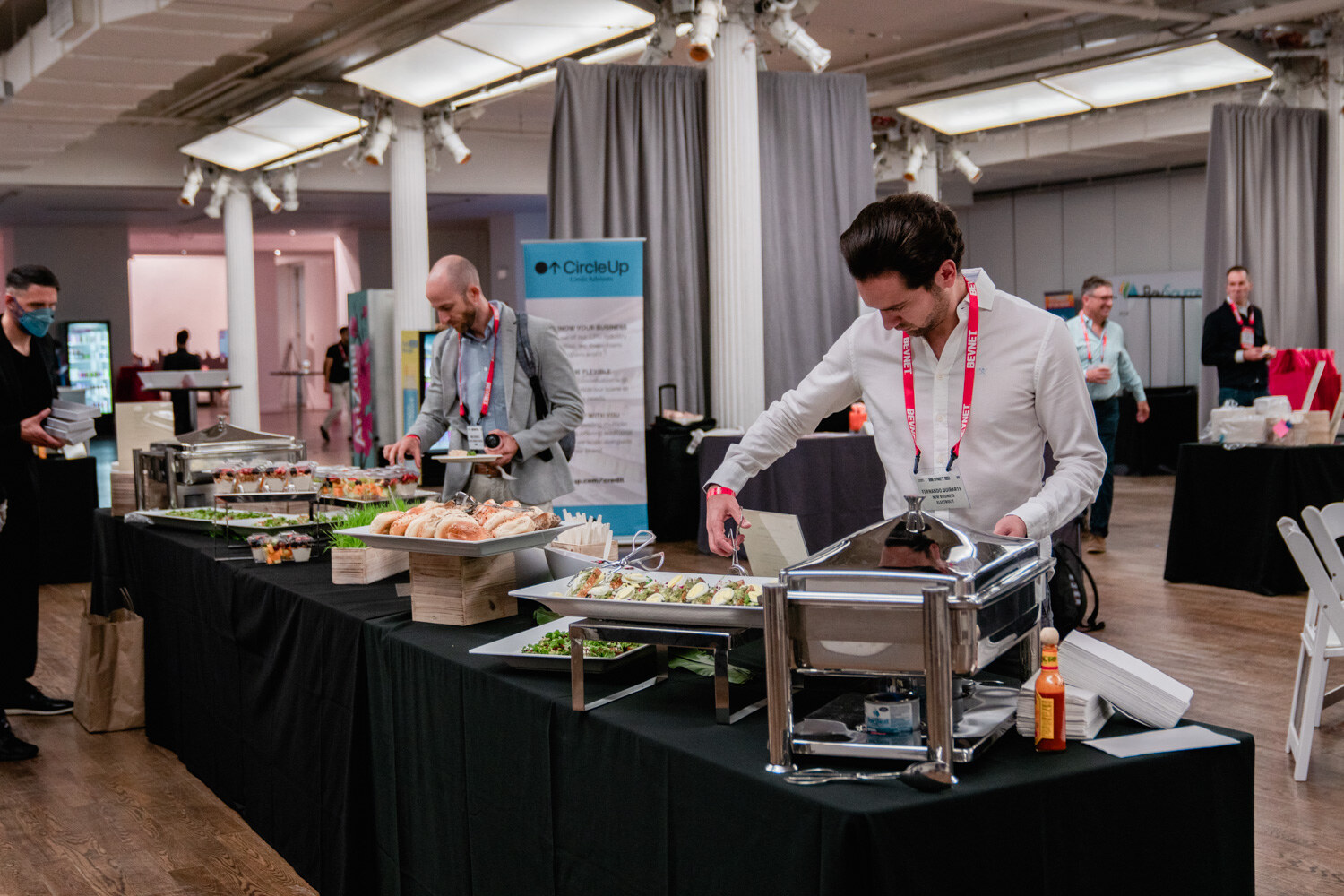 Two men in lanyards fill plates at a long black-draped buffet table.