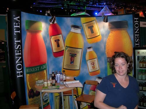 A woman with curly hair in a navy shirt sits beside a small table in a booth with large Honest Tea and Honest Ade bottle graphics and a refrigerated cooler at the right.