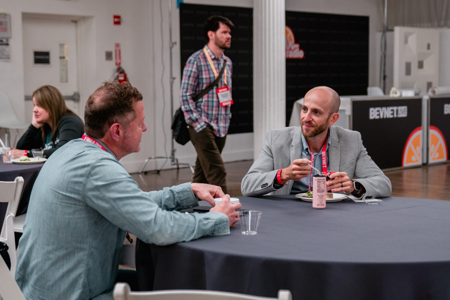 A man in a gray blazer and a man in a teal shirt sit at a round table with plates and cups, a Taste Radio stage in the background.
