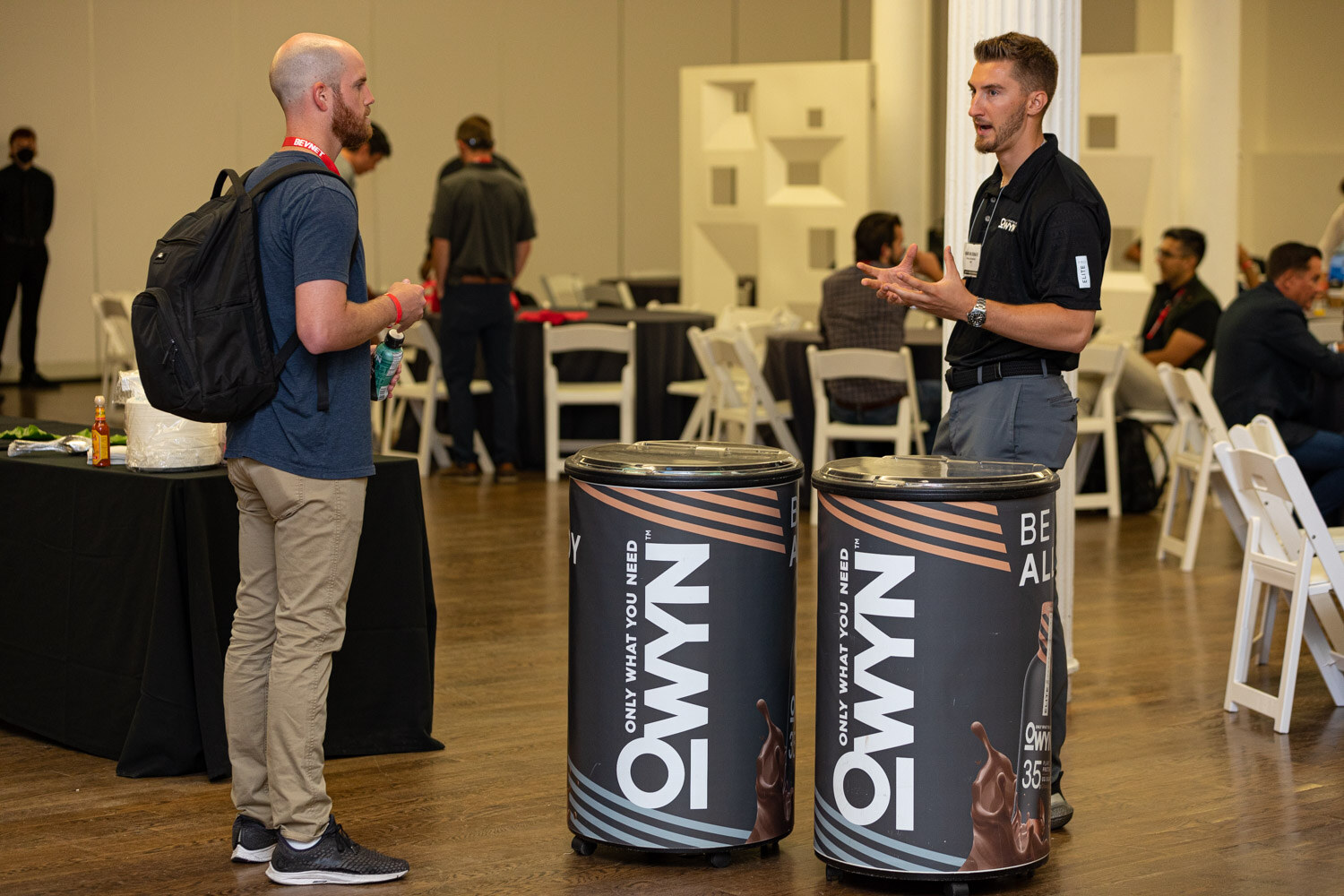 Two black cylindrical coolers branded Owyn stand on a wood floor as a man in a backpack talks to a man in a black shirt.