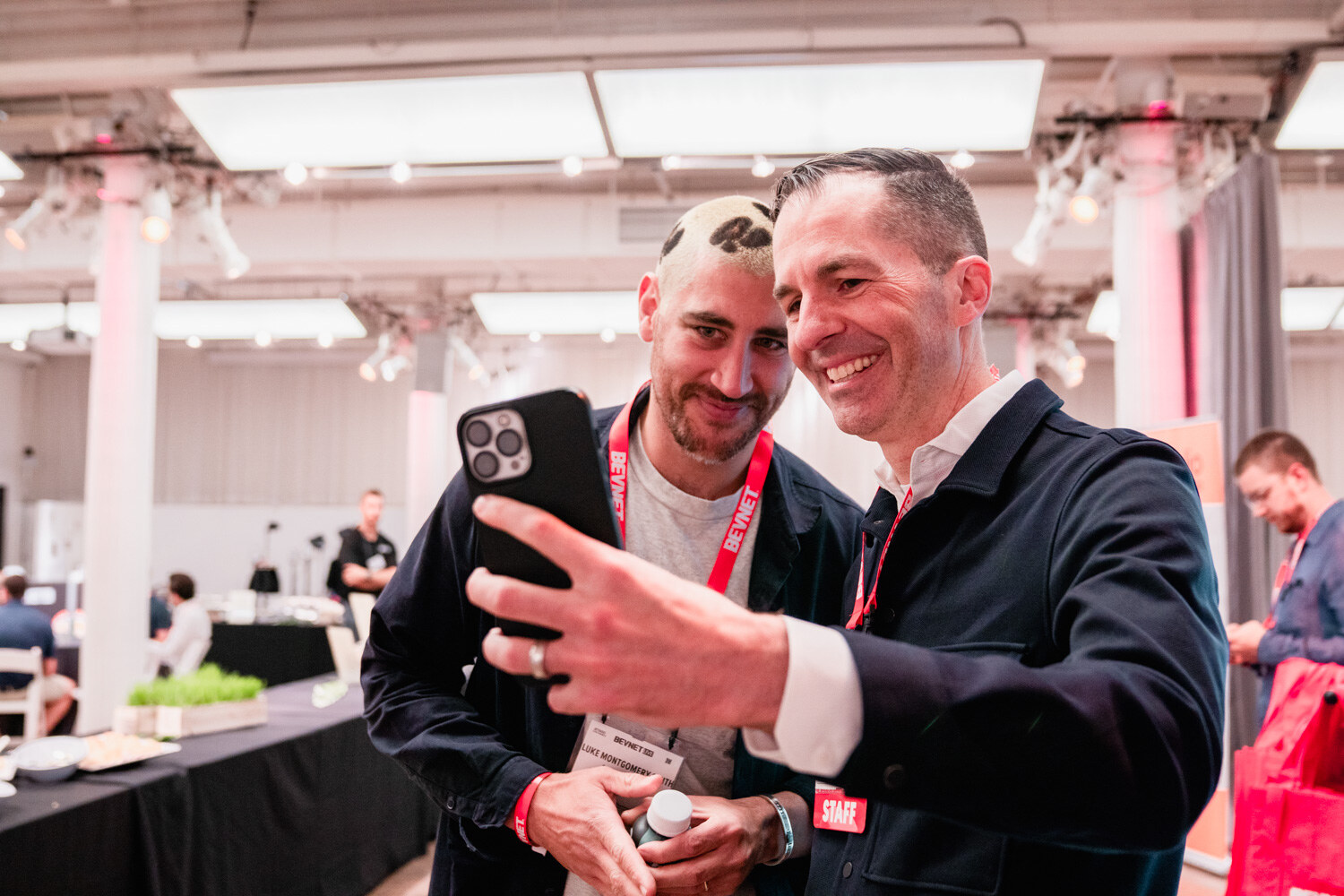 Two men in lanyards pose for a selfie, one holding a phone toward the camera, in a bright hall with tables in the background.