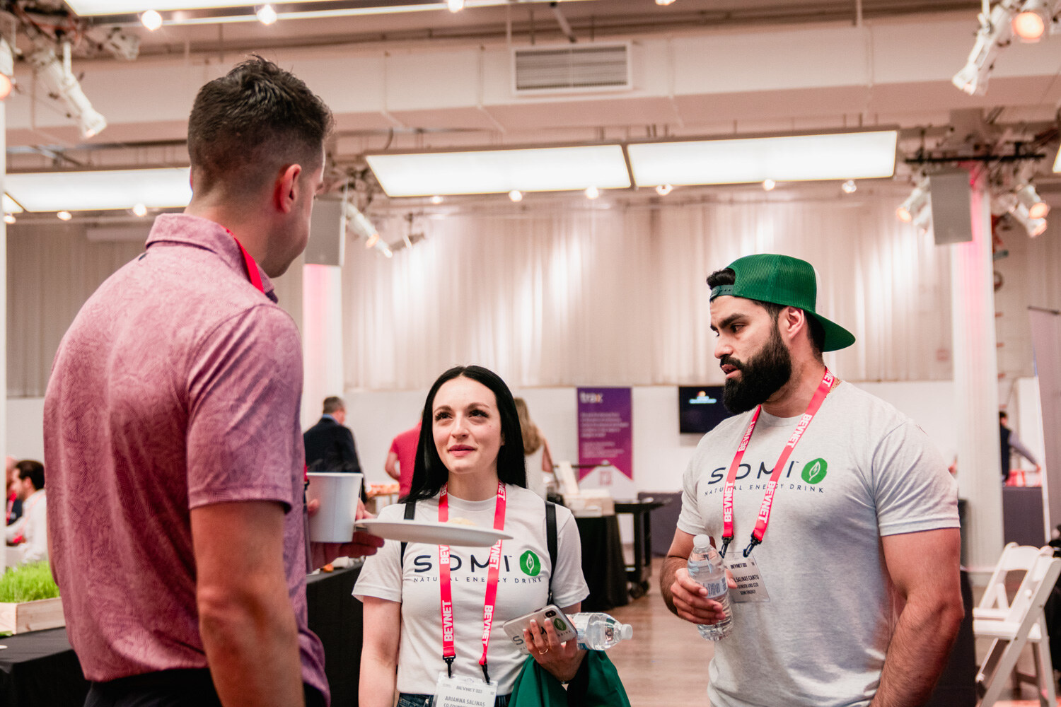 A woman and a bearded man in white Somi shirts with lanyards talk to a man in a pink polo.