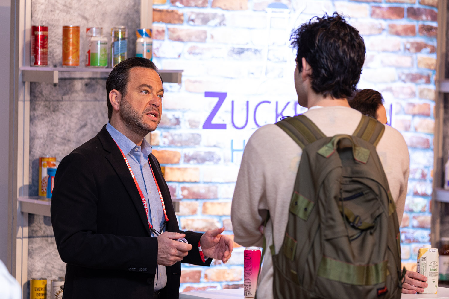 A man in a black blazer talks to a man wearing a green backpack in front of a shelf of canned and bottled drinks.
