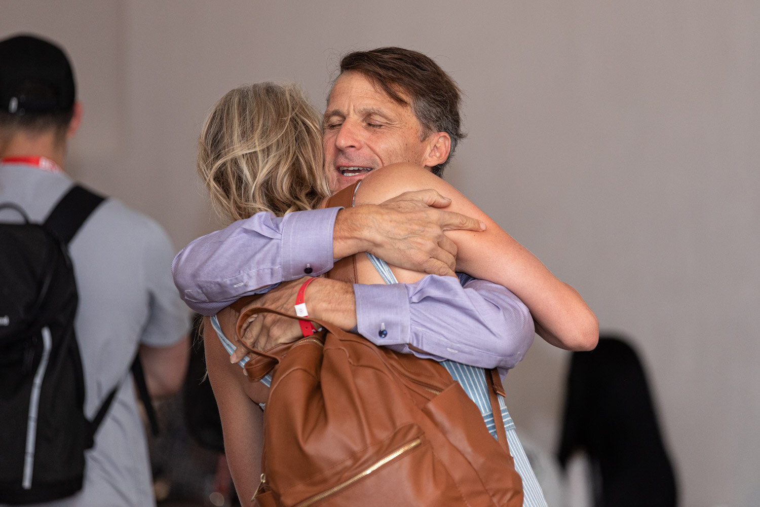 A man in a lilac shirt hugs a woman carrying a brown leather bag.