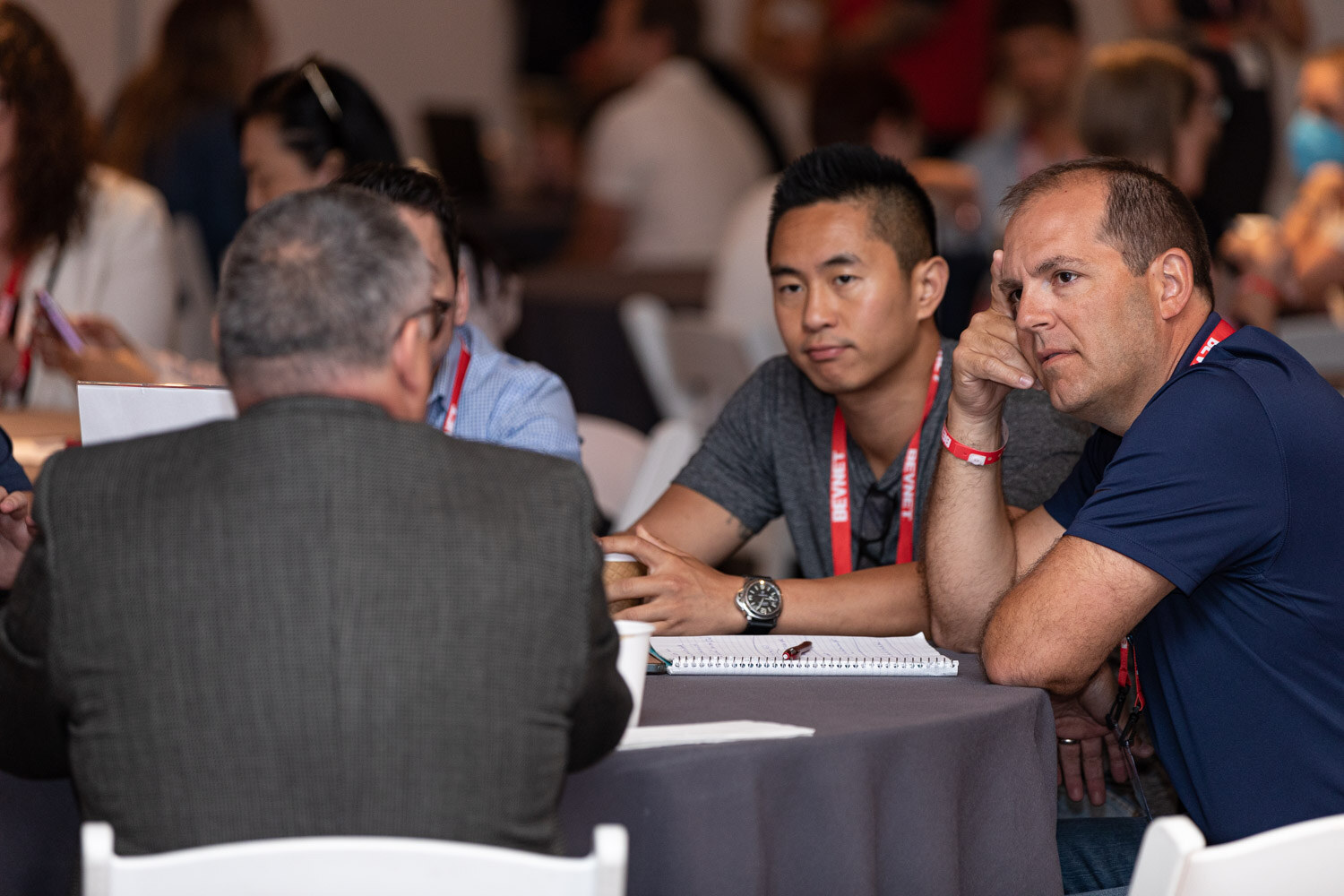 A man in a navy polo and a man in a gray shirt with lanyards sit at a round table, listening to a person on the left.