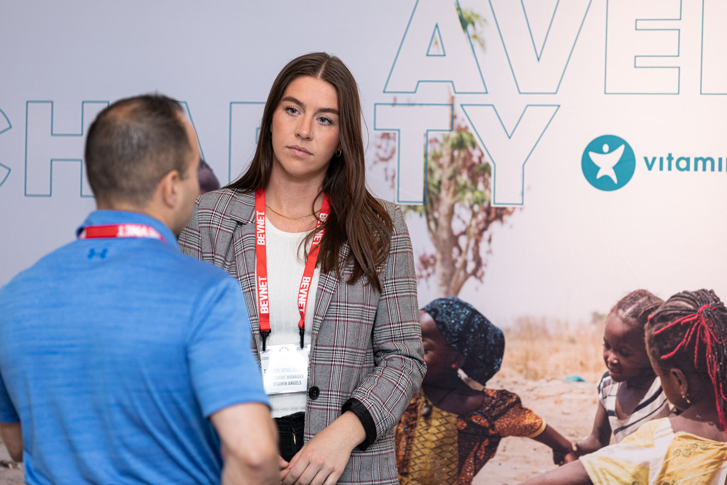 A woman in a gray blazer with a lanyard looks at someone in a blue polo in front of a wall graphic with children and the words Charity.