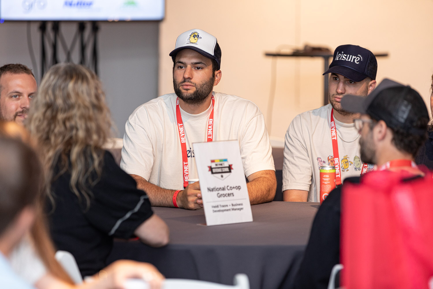 Men in cream t-shirts and caps sit at a round table with a National Co-op Grocers table card and a red lanyard strap.