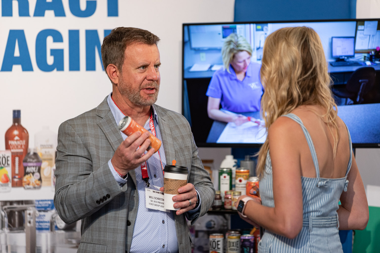 A man with a coffee cup and a can speaks to a woman in a striped dress in front of a screen and shelves of drinks.