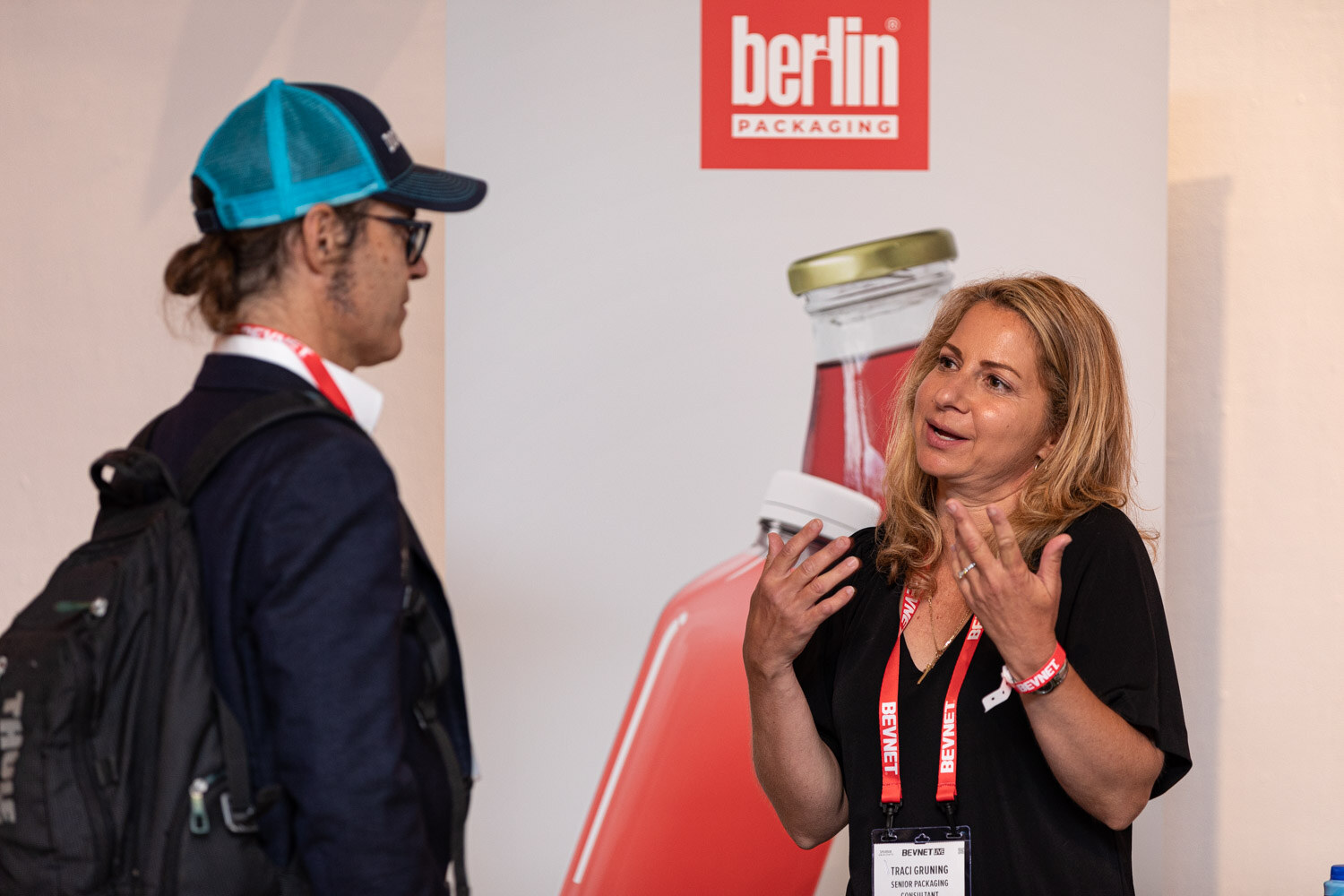 A woman in a black top and a lanyard gestures while talking to a man in a teal cap in front of a Berlin Packaging banner.