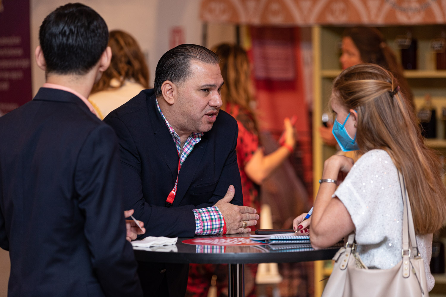 A man in a dark blazer talks to a woman in a face mask and a white top at a high-top table, with notes in front of them.
