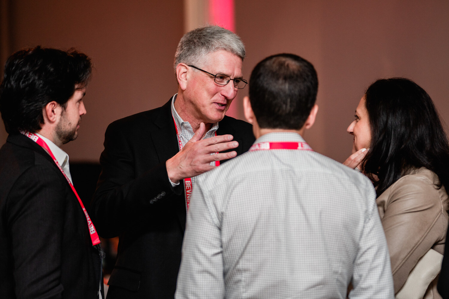 A gray-haired man with glasses gestures while talking to a woman and two men in lanyards.