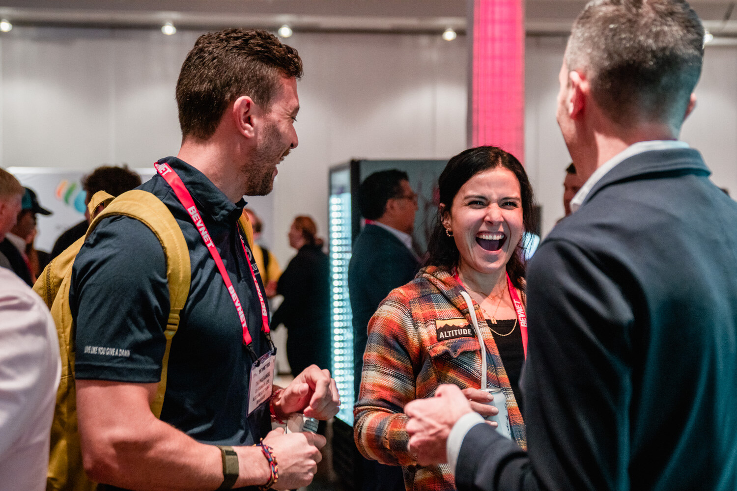 A woman in a plaid Altitude jacket laughs with two men in lanyards in the hall.
