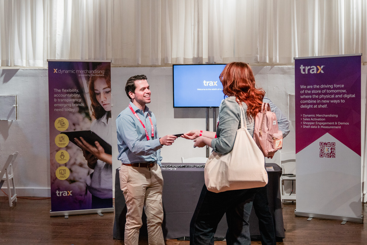 A man in a blue shirt hands a card to a woman with a tote bag in front of purple Trax and Dynamic Merchandising banners.